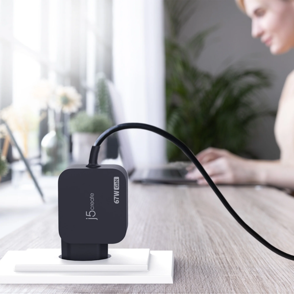 A black jbl wireless GT-Y60 60W charger plugged into a white wall socket on a wooden desk, with a blurred woman working in the background.