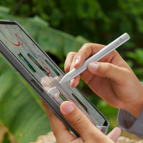 A close-up of a person's hands using a white stylus on a tablet screen, with a blurred green outdoor background.
