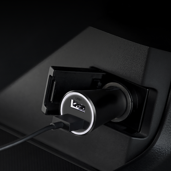 A close-up of a black USB car charger plugged into a car's cigarette lighter socket, with a USB cable inserted. The charger features a silver ring around the USB port and a circular 'USB 3.