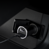 A close-up of a black USB car charger plugged into a car's cigarette lighter socket, with a USB cable inserted. The charger features a silver ring around the USB port and a circular 'USB 3.