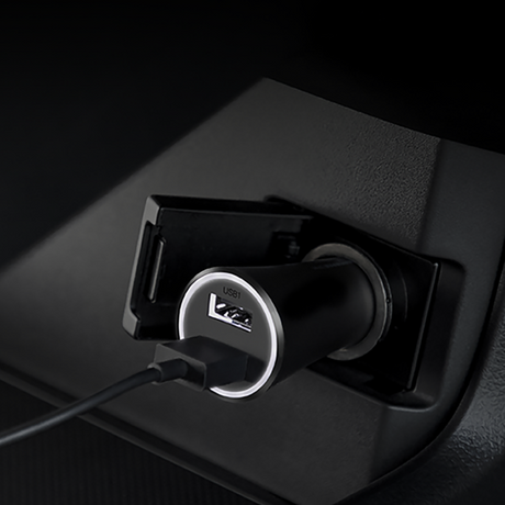 A close-up of a black USB car charger plugged into a car's cigarette lighter socket, with a USB cable inserted. The charger features a silver ring around the USB port and a circular 'USB 3.