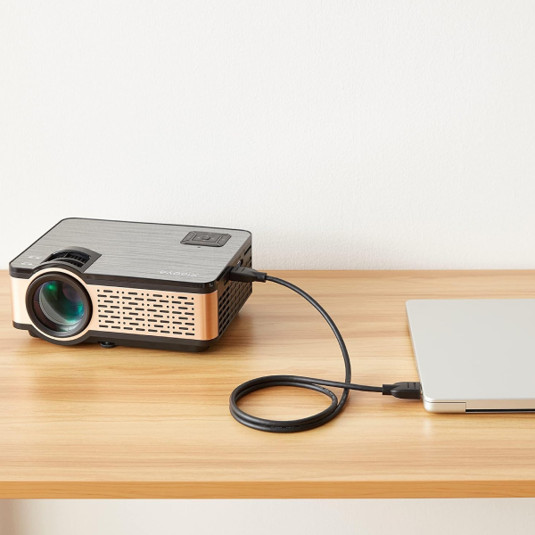 A compact black and metallic projector connected via a black cable to a silver laptop, placed on a light wood desk against a plain off-white wall.