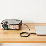 A compact black and metallic projector connected via a black cable to a silver laptop, placed on a light wood desk against a plain off-white wall.