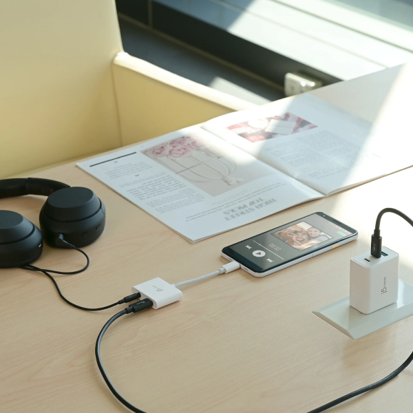 A smartphone sits on a light wood desk connected to a white audio adapter, with black over-ear headphones on the left and a white wall charger on the right. An open magazine lies behind the phone in a bright indoor setting.