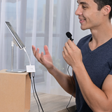 A man wearing a navy blue t-shirt is shown in a lifestyle setting, holding a small black lavalier microphone and gesturing towards a white USB adapter connected to a tablet on a silver stand and a cardboard box.