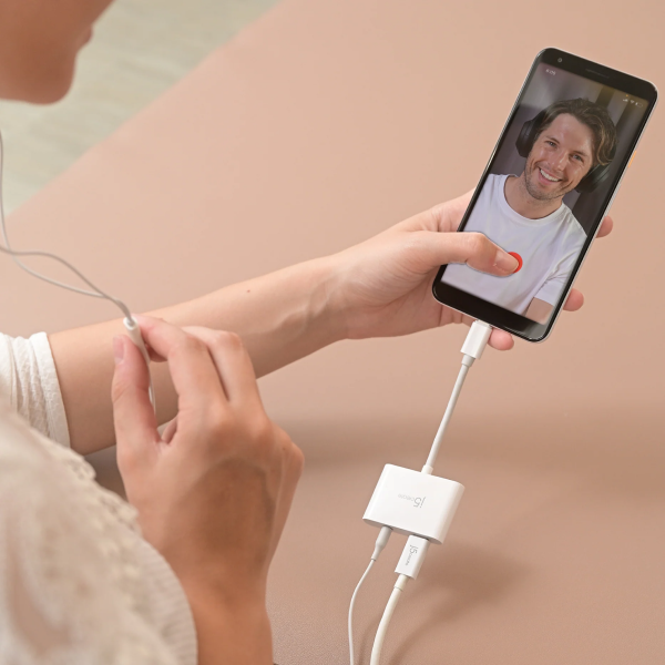 A person holds a smartphone connected to a white USB dongle and wired earbud, with a video call visible on the phone screen against a soft beige background.