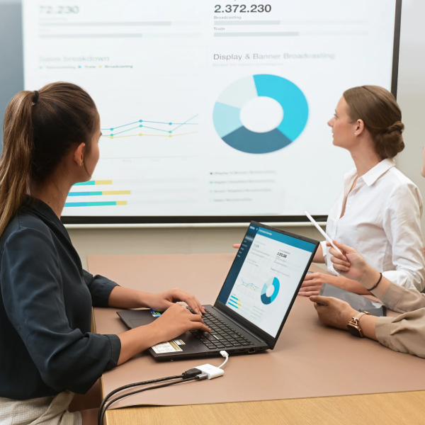 Two women working together at a desk with a laptop, discussing data displayed on a screen.