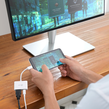 Person using a smartphone with a USB hub connected to a computer monitor on a desk.