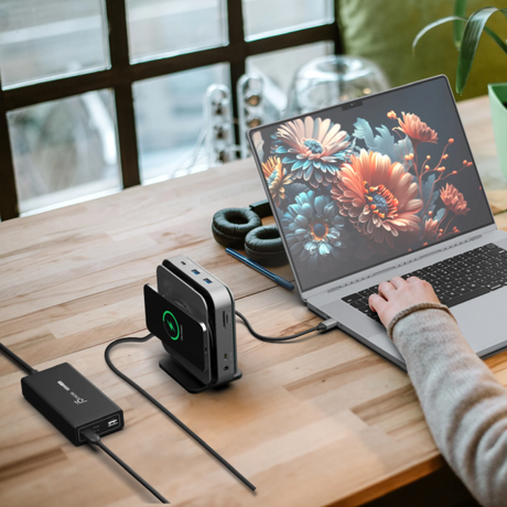 A desktop setup shows a laptop on a wooden table with a person's arm resting on it, alongside a black external hub/device with a green charging light and a black cable. Headphones sit behind the device, and a window and indoor decor are visible in the background.