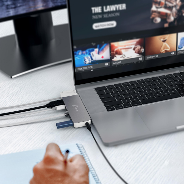 Laptop with a USB hub attached on a desk, with a person's hand holding a pen.