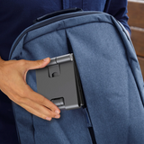 A close-up of a person's hand sliding a dark gray, foldable tablet holder into the front pocket of a blue backpack.