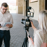 A woman films a man with a DSLR camera on a tripod in an outdoor walkway, while the man holds a tablet. The camera has an external monitor showing the video feed.