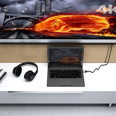 j5create JDC52 Premium High-Speed HDMI®™ Cable with Etherne  (6.6ft)  A 4K wired projector screen mounted on a wall above a white desk, displaying a glowing orange car graphic. A laptop, black headphones, and a small notebook are arranged on the desk below.
