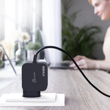 A black jbl wireless GT-Y60 60W charger plugged into a white wall socket on a wooden desk, with a blurred woman working in the background.